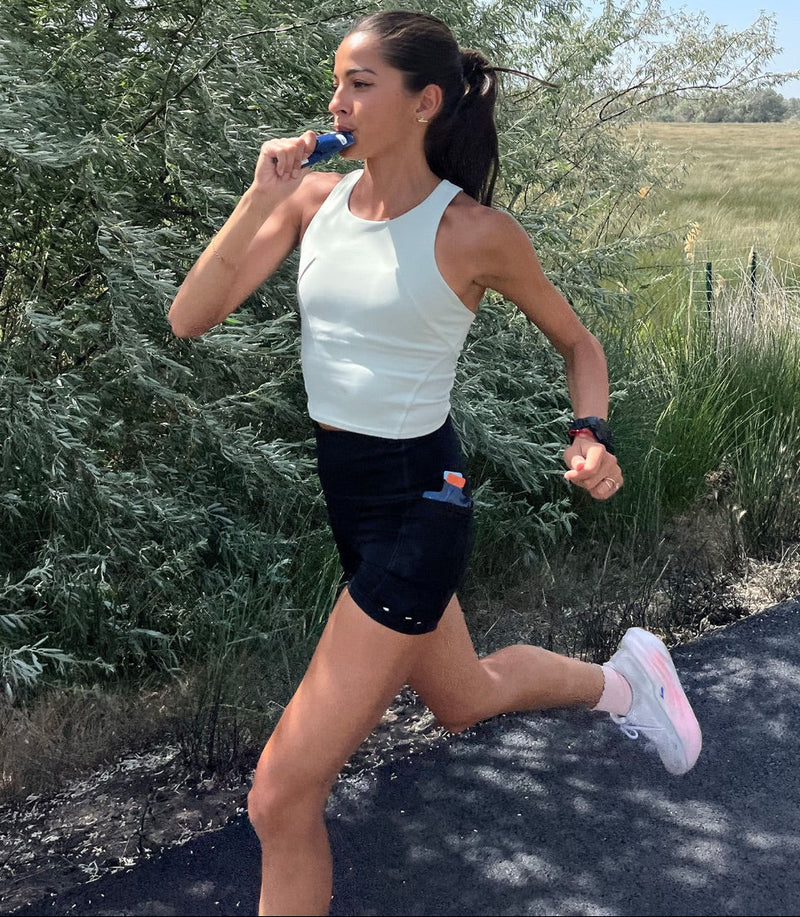 A woman running outdoors on a paved path wearing a white athletic tank top, black running shorts, and white running shoes, with green bushes and a field in the background.